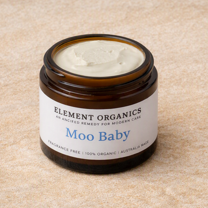 An amber glass jar of Element Organics 'Moo Baby' cream with a white label, set on a textured beige surface.