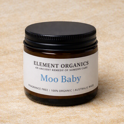 An amber glass jar candle with a black screw-on lid and a white label reading 'ELEMENT ORGANICS' and 'Moo Baby' on a beige textured background.
